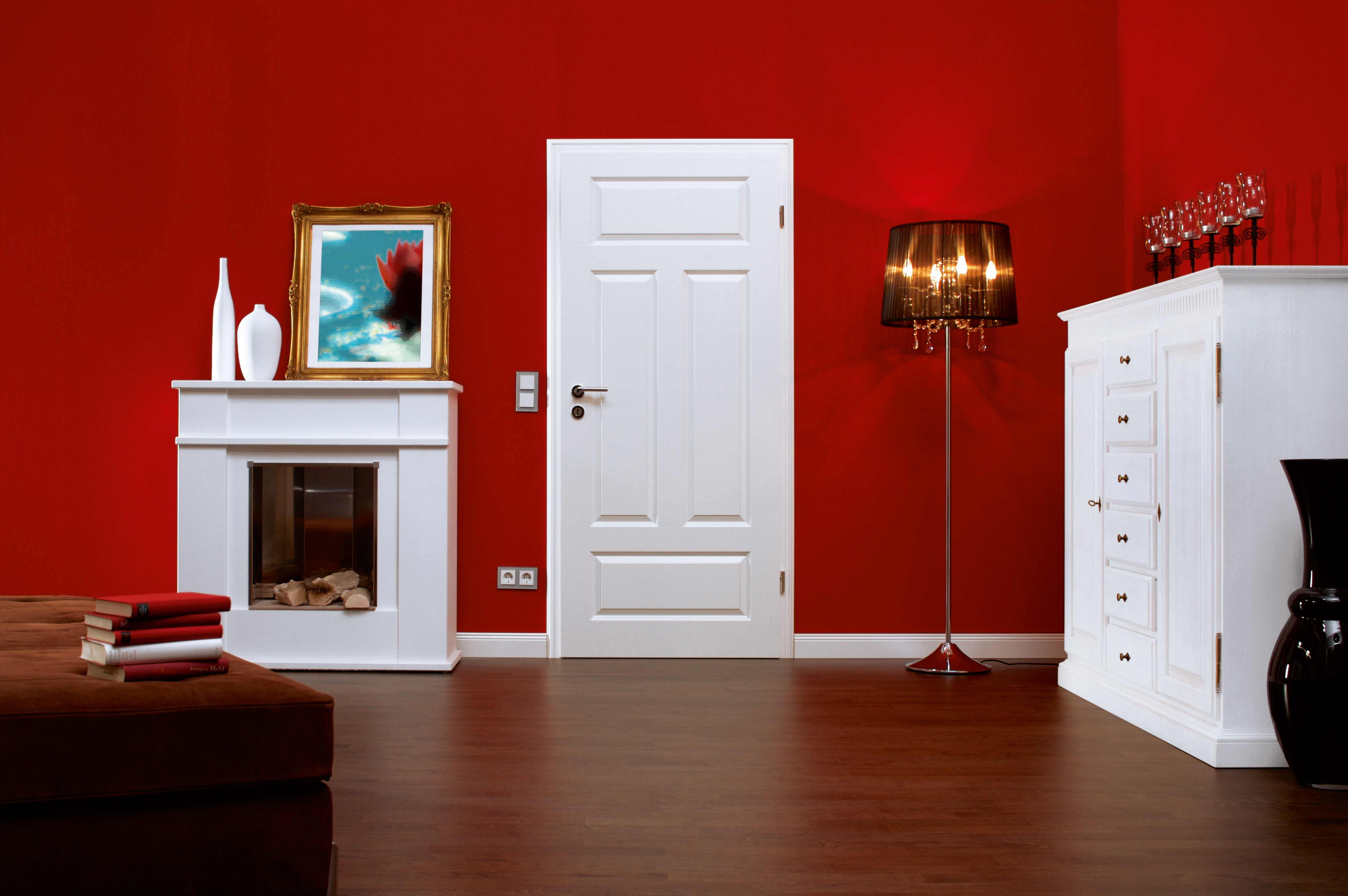 Ein rot gestrichener, großer Raum mit einer weißen Tür, einem weißen Kamin, einem weißen Schrank, einem Gemälde mit Goldrahmen und einer Stehlampe