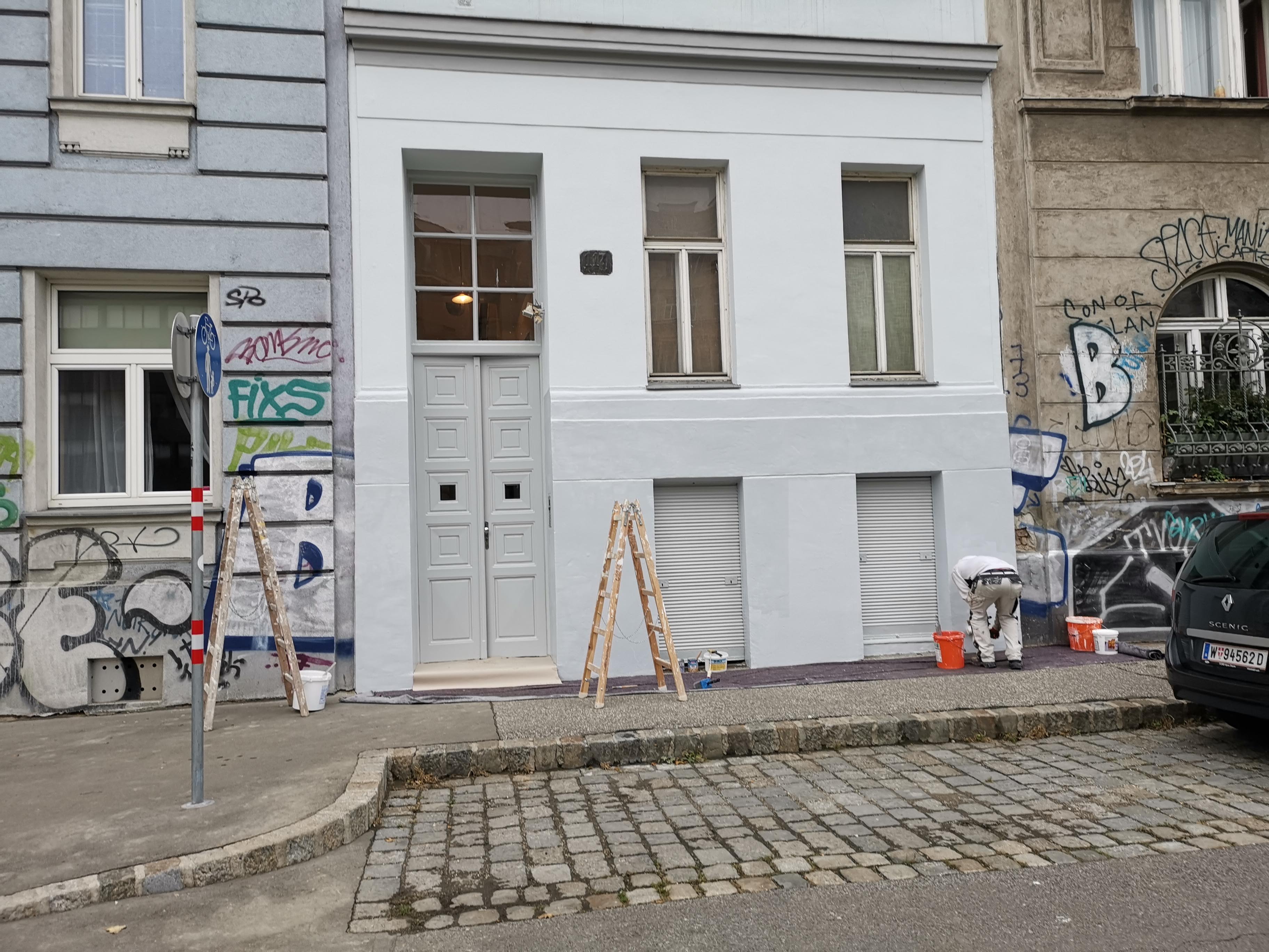An exterior photo of the front of a grey painted building in a street. A man is at work with a paint brush