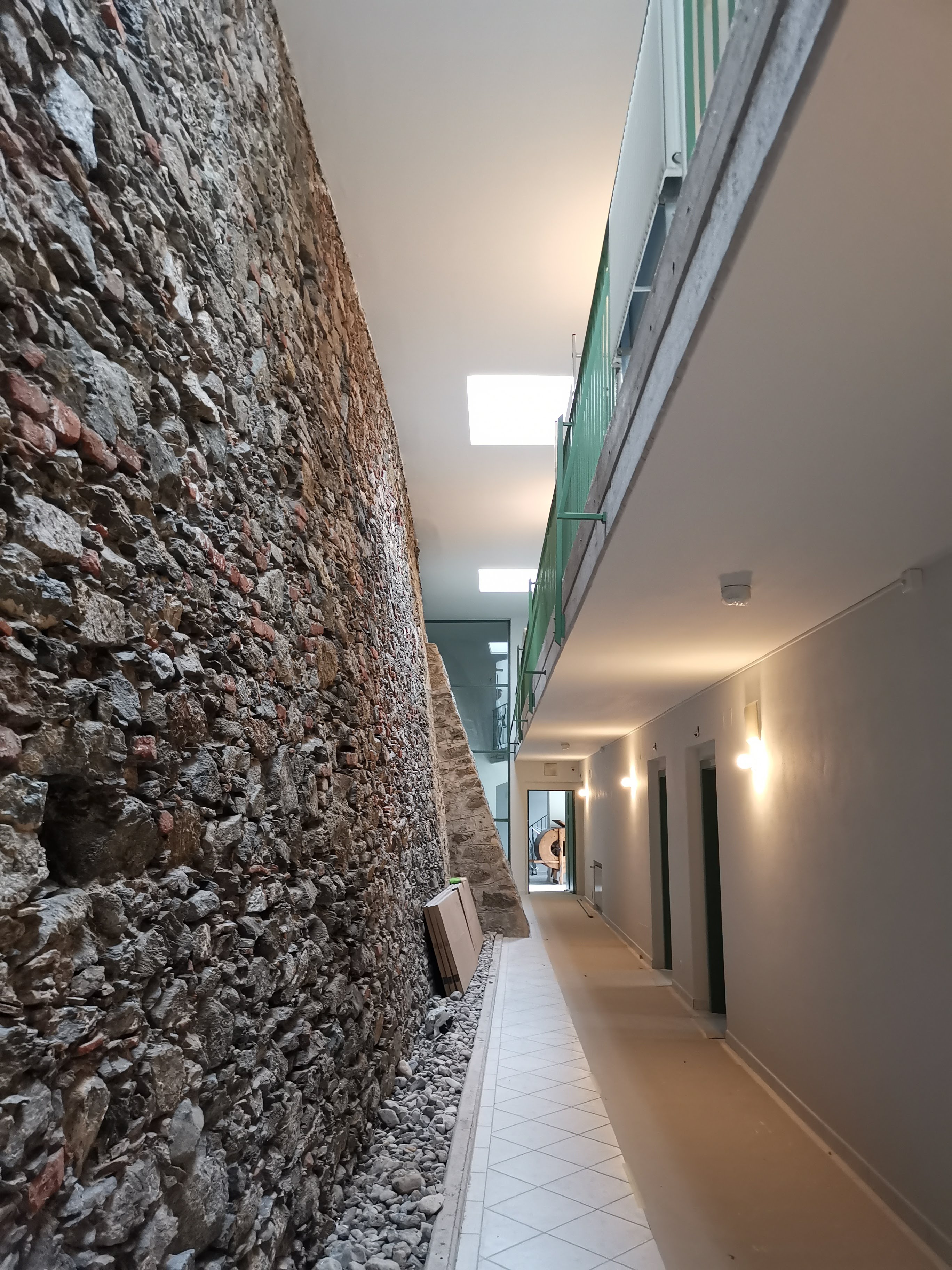 Eine Innenansicht eines Korridors. Auf der linken Seite verläuft eine alte Steinmauer, die rechte Seite ist hoch und modern